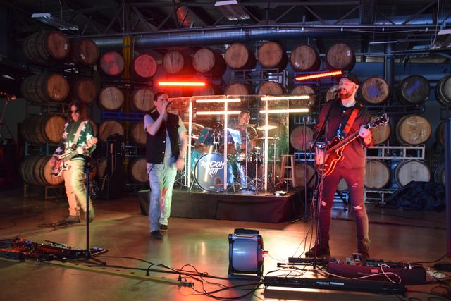 Wide shot of Random Riot mid-song, drummer lit behind the shield and the band spread across the stage
